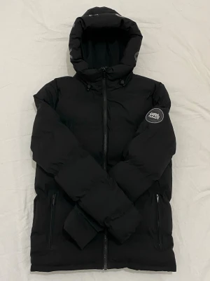 Svart pufferjacka från Supply and Demand - Prisen kan diskuteras. Snygg svart pufferjacka från Supply And Demand med huva och dragkedja framtill. Jackan har två fickor med dragkedja och en patch med logga på ena ärmen. Perfekt för kalla vinterdagar tack vare det vadderade materialet. Den är bara använt två gånger.