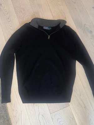 Ralph Lauren half zip - Klassisk svart tröja från Polo Ralph Lauren med halv dragkedja och ribbad krage i grått. Långärmad modell i mjukt material, perfekt för lager på lager. Enkel och stilren design som funkar till det mesta.