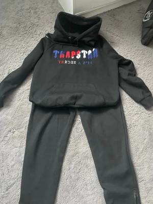 Svart set från Trapstar med tryck - Säljer ett svart set från Trapstar med hoodie och matchande byxor. Hoodien har färgglatt tryck i blått, rött och vitt samt texten 'IT'S A SECRET'. Byxorna har resår och dragsko i midjan. Materialet är mjukt och bekvämt, perfekt för en avslappnad look.