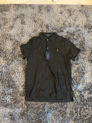 Mörkgrå pikétröja från Ralph Lauren - Snygg mörkgrå pikétröja från Polo Ralph Lauren med klassisk krage och två knappar framtill. Tr pipeline har korta ärmar och den ikoniska broderade loggan på bröstet. Perfekt till jeans eller shorts för en clean look.