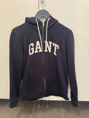 GANT full zip hoodie med huva - Snygg full zip hoodie från GANT med stor logga framtill. Mörkblå med vita dragsnören i huvan och kängurufickor. Säljes för 100kr. 