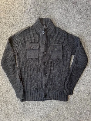Mörkgrå kabelstickad kofta - Säljer en mörkgrå kabelstickad kofta med knappar framtill och två bröstfickor med lock och knapp. Koftan har hög krage, ribbade muddar och axelvaddar. Perfekt för dig som gillar en chunky och stilren look.
