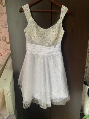 Vit glittrig klänning med paljetter till fest och balen  - Använd en gång. Vit klänning med paljetter och pärlor på överdelen. Klänningen har en markerad midja med ett brett, veckat band. Kjoldelen består av flera lager tyll med paljettdetaljer, vilket ger en voluminös och festlig look. Klänningen har en dragkedja i sidan för enkel påklädning och inbyggda kupor i bystdelen. 