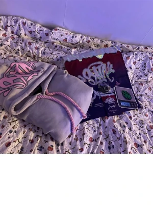 Ljusgrå hoodie med rosa detaljer - Säljer en ljusgrå hoodie från SANTA med stor rosa logga på huvan och matchande rosa dragsnören. Hoodien har en klassisk känguruficka framtill och är gjord i mjukt material, perfekt för chill dagar. Oversize känsla och streetwear-vibe.