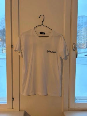 Vit t-shirt från Palm Angels - Vit t-shirt från Palm Angels med svart logotyptryck på bröstet. Klassisk rund halsringning och korta ärmar. Perfekt basic-plagg med clean design och skön passform. Det står Palm angels där bak på ryggen med