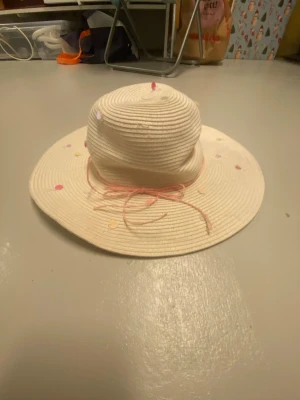 Vit solhatt med färgglada prickar - Säljer en söt vit solhatt i flätad strådesign med bred brätte. Hatten har färgglada konfettiprickar och ett tunt rosa band knutet runt kullen. Perfekt för soliga dagar och ger en lekfull touch till din outfit.
