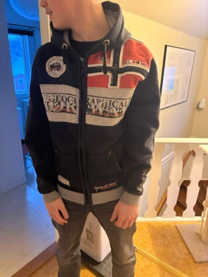 Svart hoodie Geographical Norway - Snygg svart hoodie från Geographical Norway med dragkedja, huva och stora tryck på både fram- och baksidan. Perfekt för dig som gillar streetwear och tydliga prints.