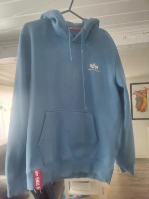 Blå hoodie Alpha Industries  - Snygg blå hoodie från Alpha Industries med vit logga på bröstet och stor känguruficka framtill. Huva med dragsko och ribbade muddar. Röd Frank Dandy-tag vid nederkanten. Perfekt för en chill och avslappnad stil. Kan finnas någon liten fläck men är ny 