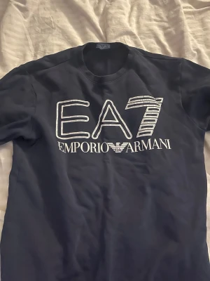  EA7 Emporio Armani tröja - Snygg marinblå t-shirt från EA7 Emporio Armani med stort vitt tryck på bröstet. Klassisk passform och rund halsringning. Perfekt för dig som gillar stilrena loggor och exklusiv streetwear.