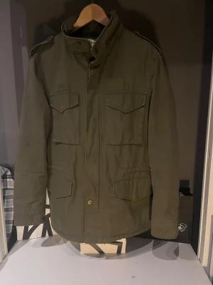 Olivgrön vindjacka från Dobber - Stilren olivgrön vindjacka från Dobber med fyra framfickor och hög krage. Jackan har axelklaffar och stängs med dragkedja och tryckknappar. Tillverkad i slitstarkt material, perfekt för blåsiga dagar.