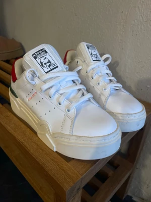 Adidas Stan Smith vita sneakers - Fräscha vita Adidas Stan Smith sneakers i läder med gröna och röda detaljer på hälen. Klassisk låg modell med perforerade ränder på sidan och Stan Smith-logga på plösen. Sköna och stilrena skor som passar till det mesta.