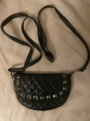 Svart quiltad axelväska med nitar - Snygg svart axelväska i quiltad design med silverfärgade nitar på framsidan. Väskan har lång justerbar axelrem. Perfekt för dig som gillar en edgy och trendig stil.
