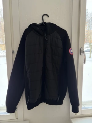 Svart quiltad jacka från Canada Goose - Snygg svart jacka från Canada Goose med quiltad front, huva och dragkedja. Jackan har lång ärm och två fickor med dragkedja. Märkespatch på ärmen ger en exklusiv touch. Perfekt för kyliga dagar.