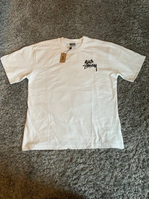 Vit Stüssy t-shirt med tryck - Säljer en vit t-shirt från Stüssy med svart logga på bröstet och stort grafiskt tryck på ryggen med en ängel och en biljardboll. Klassisk passform och rund hals.