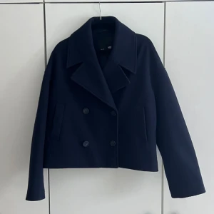 Mörkblå kort kappa - Snygg mörkblå kappa med dubbelknäppning och bred krage. Modellen är kort och har raka ärmar samt två diskreta fickor framtill. Perfekt för dig som vill ha en stilren och modern look. Materialet ser ut att vara ullblandning.