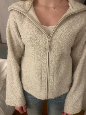Krämvit teddyjacka med dragkedja - aldrig använd, Mysig krämvit teddyjacka med hel dragkedja framtill och hög krage. Jackan har en fluffig, fårskinnsinspirerad yta och två sidofickor. Perfekt för att hålla värmen under kyliga dagar och ger en avslappnad, trendig look.