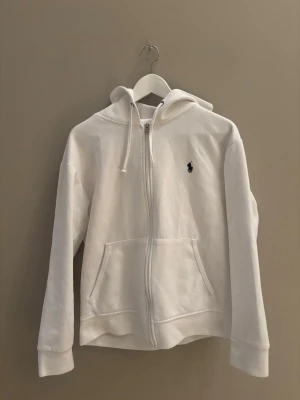 Vit zip hoodie från Polo Ralph Lauren - Klassisk vit hoodie med dragkedja från Polo Ralph Lauren. Hoodien har huva med dragsko, två fickor framtill och den ikoniska lilla loggan broderad på bröstet. Tillverkad i mjukt bomullsmaterial som känns skönt mot huden. Perfekt för en clean och stilren look.