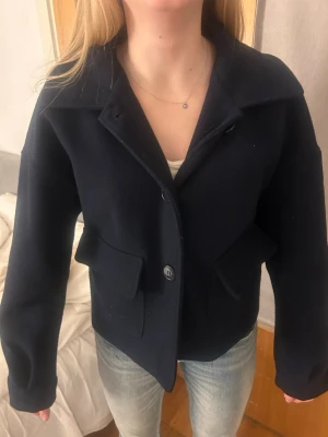 Mörkblå kappa  - Stilren mörkblå kappa med bred krage och stora framfickor. Kappan har knäppning framtill med två knappar och är tillverkad i ett tjockare material som ser ut att vara ull. Passar perfekt till jeans för en clean look.