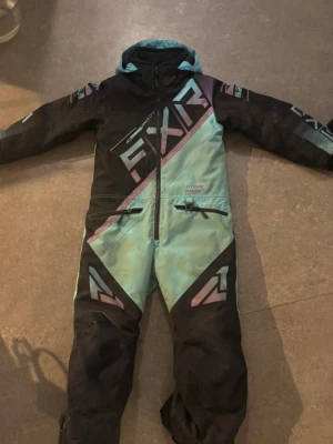 barn FXR overall i svart och turkos - Säljer en snygg overall från FXR i svart, turkos och rosa med stora logotyper och dragkedja framtill. Overallen har huva, två fickor med dragkedja och coola grafiska detaljer. Perfekt för dig som vill sticka ut i vinterlandskapet. Strl 6, skulle säga att det passar 116-134 