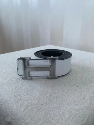 Vitt skärp med H-spänne - Snyggt vitt skärp med ett stort H-format spänne i silverfärg. Bältet har en stilren design och är tillverkat i skinn. Perfekt accessoar för att lyfta din outfit med en exklusiv touch.