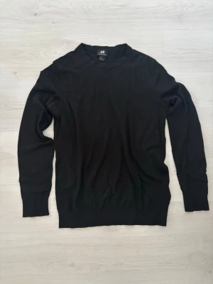 Svart stickad tröja från H&M - Klassisk svart stickad tröja från H&M i merinoullsblandning. Tröjan har rund halsringning, långa ärmar och ribbade muddar vid ärmslut och nederkant. Perfekt basplagg för lager på lager och enkel att matcha med olika stilar.