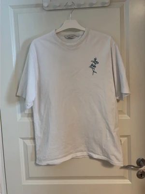 Vit t-shirt Casual Friday med tryck - Vit t-shirt från Casual Friday med mörkblått blommigt tryck på bröstet och större tryck på ryggen med texten 'Casual Friday Aesthetics Club'. Klassisk passform och rund halsringning. Perfekt för en clean och stilren look.