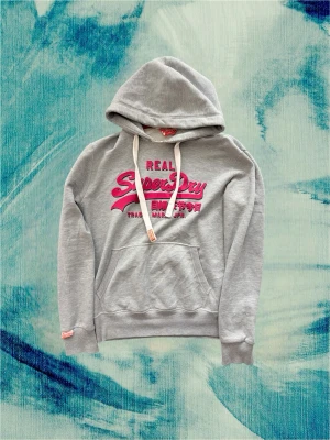 Grå Superdry hoodie med rött tryck - Sjukt fet hoodie från Superdry! Passar dig som är mellan 187-193. Sjukt fett med de röda detaljerna, passar väldigt bra till ett par mörka jeans. Hör av dig! Jag är 190 och väger 80 kg för referens
