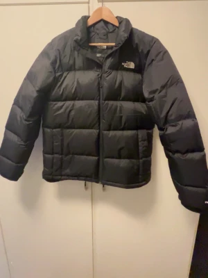 Svart pufferjacka från The North Face - Säljer en svart pufferjacka från The North Face med klassisk logga på bröstet. Jackan har en hög krage, dragkedja framtill och quiltad design. Perfekt för kalla vinterdagar och tillverkad i ett slitstarkt material.