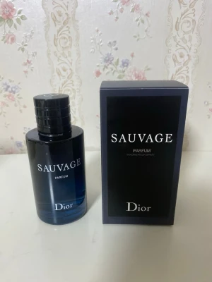 Dior Sauvage Parfum 100ml - Dior Sauvage Parfum är en lyxig herrdoft i en elegant mörkblå glasflaska med svart lock. Flaskan har en stilren och modern design med tydlig logotyp. Parfymen levereras i en matchande svart kartong med vit text.
