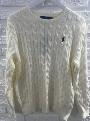 Vit kabelstickad tröja Polo Ralph Lauren - Kabelstickad vit tröja från Polo Ralph Lauren med klassisk marinblå logga broderad på bröstet. Tröjan har rund halsringning, långa ärmar och ribbade muddar. Perfekt för lager på lager och stilrena outfits.