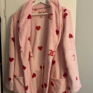  Rosa Juicy Couture morgonrock med röda hjärtan - Supermjuk rosa morgonrock från Juicy Couture med röda hjärtan och JC-loggor. Har två stora fickor framtill, bälte i midjan och väldigt mysig. Perfekt för chilliga morgnar eller kvällar hemma.
