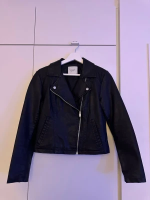 Svart skinnjacka från Jacqueline de Yong  - Snygg svart skinnjacka med klassisk bikerdesign, sned dragkedja och breda slag. Jackan har två fickor framtill och stilrena detaljer i silver. Perfekt för att lyfta din outfit med en cool touch.