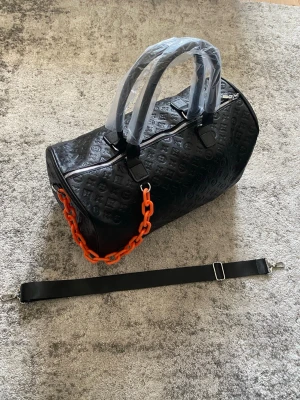 Svart Duffle bag med mönster - Riktigt snygg och stilren väska i helt nytt skick, aldrig använd. Kedjan är endast på för stilens skull, kan tas bort och bytas ut mot remmen i bild 1. Hör av dig vid funderingar så löser vi!