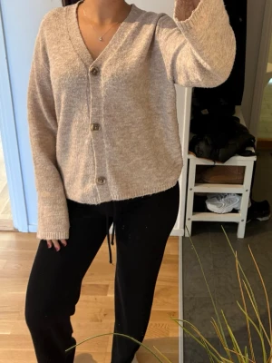 Beige kofta med knappar - Mysig beige kofta med v-ringning och stora knappar framtill. Koftan har lång ärm och är tillverkad i ett mjukt, stickat material som ger en avslappnad look. Perfekt att slänga på sig över en t-shirt eller linne.