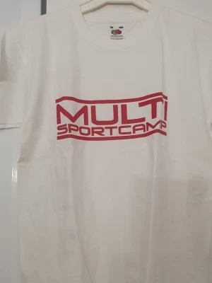 T-shirt - Vit t-shirt från Fruit of the Loom med röd MULTI SPORTCAMP-tryck både fram och bak. Klassisk rund hals och korta ärmar. Tillverkad i mjuk bomull som känns skön mot huden. Perfekt för dig som gillar sportig och enkel stil.