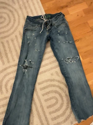 Blå slitna jeans med hål - Säljer ett par blå jeans med flera slitningar och stora hål på benen. Jeansen har rak passform, snörning i midjan istället för vanlig knapp och dragkedja. Perfekta för en avslappnad och trendig streetstyle-look.