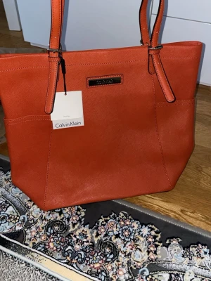 Orange handväska från Calvin Klein - Snygg orange handväska från Calvin Klein i läder med två långa axelband och stilren metallogo framtill. Väskan har en rymlig design och raka linjer, perfekt för dig som gillar en modern och trendig look. Märkets tagg sitter kvar på handtaget.