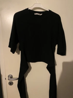 Svart topp med knyt i ryggen - Trendig svart croppad topp från Lager 157 med korta ärmar och långa knytband i midjan som ger en snygg siluett. Perfekt till jeans eller kjol för en snygg och modern look.