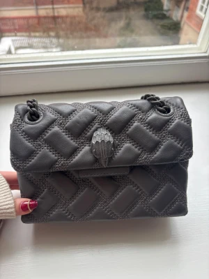 Grå quiltad axelväska Kurt Geiger - Snygg grå axelväska från Kurt Geiger med quiltat mönster och kedjeremmar i metall. Väskan har ett dekorativt silverfärgat spänne framtill och logga på baksidan. Den är i bra skick och har några små defekter på kedjan men inget som syns💕