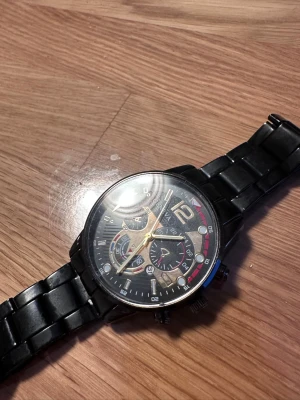 Svart analog klocka från Geneva - Stilren svart analog klocka från Geneva med metallarmband och detaljer i guld och rött på urtavlan. Klockan har flera små urtavlor, i perfekt skick och har använts väldigt sällan