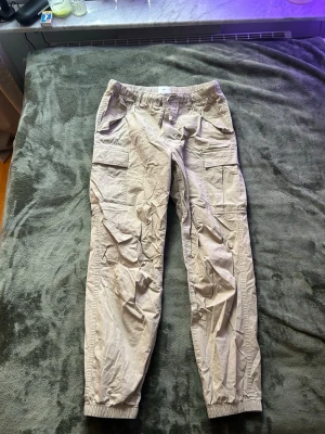 Beige cargobyxor från H&M Regular Fit - Snygga beige cargobyxor från H&M i regular fit. Byxorna har flera praktiska fickor på sidorna och resår vid bensluten för en avslappnad look. Perfekta för en chill och trendig stil. Tillverkade i ett lätt och bekvämt material.