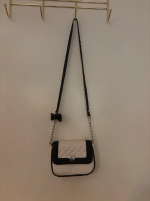 Svart och vit axelväska med rosett - Cool axelväska i svart och vitt med quiltad framsida och silverfärgad kedja. Väskan har en dekorativ svart rosett på axelbandet och ett snyggt vridlås framtill. Materialet ser ut att vara konstläder och väskan har en rektangulär form.