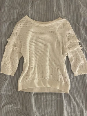 Vit stickad tröja med volangärmar - Snygg vit stickad tröja med trekvartsärmar och volangdetaljer på ärmarna. Tröjan har ett luftigt hålmönster och breda ribbade muddar vid ärmslut och nederkant. Perfekt för lager på lager och en fräsch look.❤️