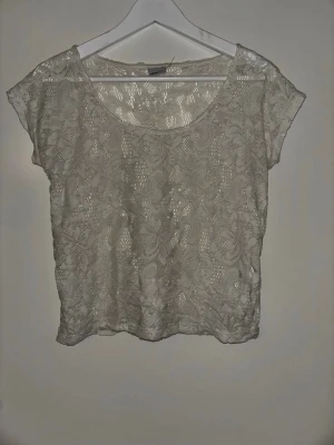 Vit spetsblus med blommönster - Söt vit blus i transparent spets med blommigt mönster. Kortärmad och croppad modell med rund halsringning. Perfekt att styla över ett linne eller bralette för en trendig look.