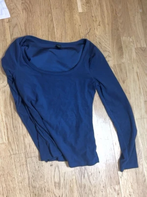 Blå långärmad topp med rund hals - En enkel blå långärmad topp med rund, djup urringning. Tillverkad i mjukt material som känns bekvämt mot huden. Perfekt att matcha med jeans eller kjol för en avslappnad look. Passar även i S