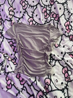 Ljuslila topp med rynkade sidor - Trendig ljuslila topp med korta ärmar och rynkade detaljer längs sidorna. Toppen har en bred, något båtformad ringning och är tillverkad i mjukt bomullsmaterial. Perfekt för dig som gillar en enkel men ändå snygg look.