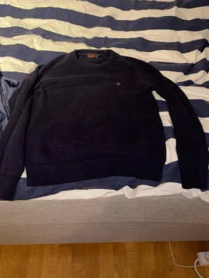 Mörkblå stickad tröja med röd detalj - Mörkblå stickad tröja med rund hals och ribbade muddar. Tröjan har en liten röd broderad detalj på bröstet. Klassisk och enkel design som passar till mycket. Materialet ser ut att vara mjukt och bekvämt.