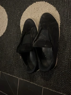 Svarta Arigato sneakers - Svarta slip-on sneakers med vit sula och minimalistisk design. Skorna har en slät ovandel utan snörning och en diskret look som passar till det mesta. Materialet ser ut att vara syntet eller mockaimitation. Perfekta för dig som gillar enkel och stilren stil.