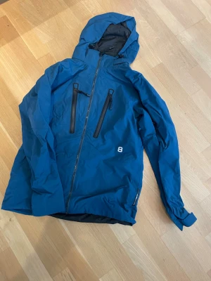 Blå vindjacka med huva från 8848 Altitude - Snygg blå vindjacka från 8848 Altitude med två svarta bröstfickor med dragkedja, hel dragkedja framtill och justerbar huva. Jackan har en sportig look och logga på bröstet och ryggen. Perfekt för blåsiga dagar och outdoor-aktiviteter.