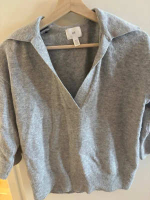 Grå tröja med krage från H&M - Snygg grå tröja från H&M med djup v-ringning och bred krage. Tröjan har ribbade muddar i nederkant och vid ärmslut, och är gjord i ett mjukt stickat material. Perfekt för en avslappnad men trendig look.